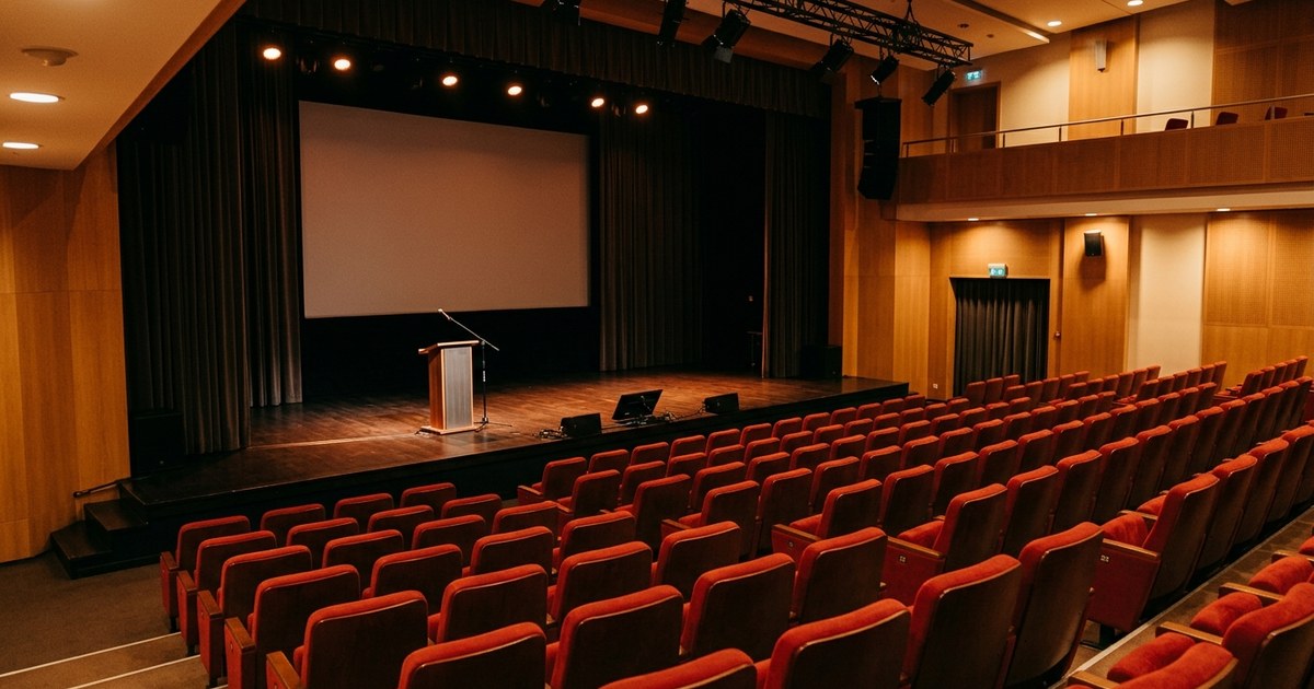 Pusta sala kinowa z rzędami czerwonych foteli, mównicą na scenie i ekranem, przed rozpoczęciem konferencji